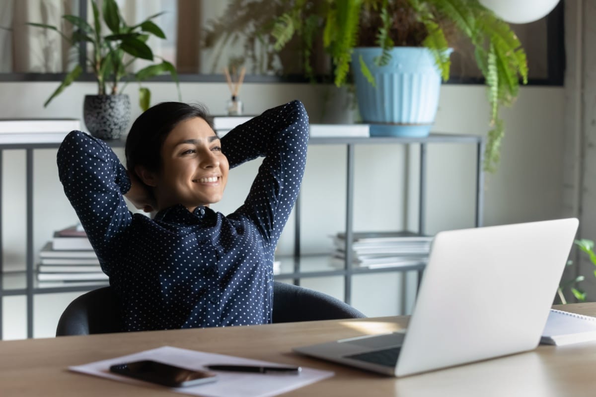 A woman in a dark blue polka-dot shirt sits at a desk in a home office, leaning back with a relaxed smile and her hands behind her head, surrounded by a laptop, phone, papers, and indoor plants—representing an anti-hustle approach to work-life balance and productivity.