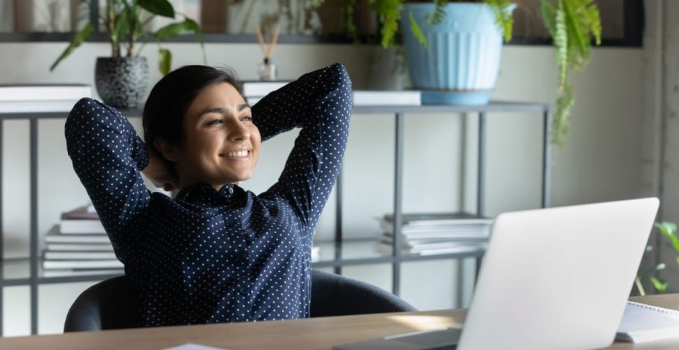 A woman in a dark blue polka-dot shirt sits at a desk in a home office, leaning back with a relaxed smile and her hands behind her head, surrounded by a laptop, phone, papers, and indoor plants—representing an anti-hustle approach to work-life balance and productivity.